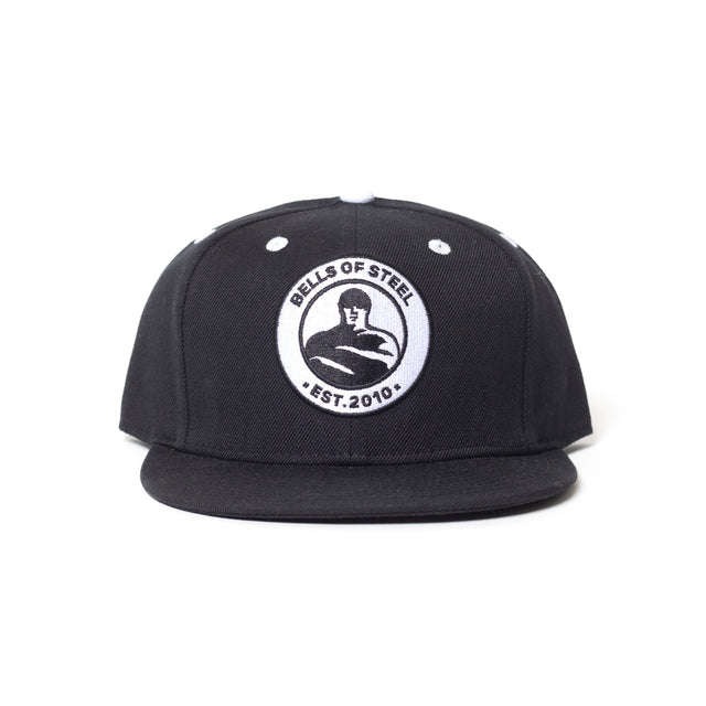 The Bells of Steel Snap Back Hat is a black flat-brim cap with a round white patch on the front, featuring "BELLS OF STEEL EST. 2010" and a kettlebell lifter—perfect for gym enthusiasts seeking style and shade.