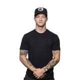 A young man with tattooed arms stands against a white background, wearing a black t-shirt, black pants, and the Bells of Steel Snap Back Hat with a white logo. He has a neutral expression and his hands are clasped in front.