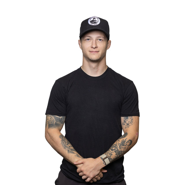 A young man with tattooed arms stands against a white background, wearing a black t-shirt, black pants, and the Bells of Steel Snap Back Hat with a white logo. He has a neutral expression and his hands are clasped in front.