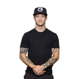 A man wearing a black t-shirt and a Bells of Steel Snap Back Hat with a round logo stands against a white background, smiling with his hands clasped in front. Tattoos are visible on both his arms.