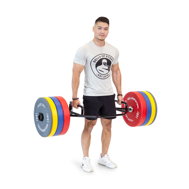 A man in a gray T-shirt and black shorts stands on a white background, confidently deadlifting with the Bells of Steel Open Trap Bar / Hex Bar, loaded with colorful weight plates. The bar incorporates rotating sleeves to ensure smooth motion as he lifts.