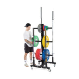 Wearing a cap, t-shirt, and shorts, a person is organizing Bells of Steel's Hydra Bumper Plate Tree Builder weights on a wheeled rack against a plain white background, highlighting the modular storage unit's sleek design.