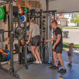 A man helps a woman working out on gym equipment in their modular home gym. A child stands nearby, fascinated by the Hydra Storage Builder from Bells of Steel, storing weights on wall racks. The open garage door reveals a driveway and house outside.