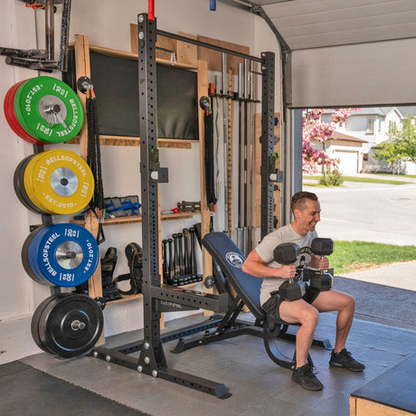 A person sits on the Bells of Steel Home Gym Builder weight bench in a garage gym, smiling with two dumbbells. Behind them are barbells, weight plates, and more equipment. Sunlight streams through the open garage door to a suburban street.