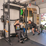 A woman uses the Bells of Steel Home Gym Builder, doing sit-ups on a weight bench while smiling at an encouraging man in a sunlit home gym with weights and power racks.