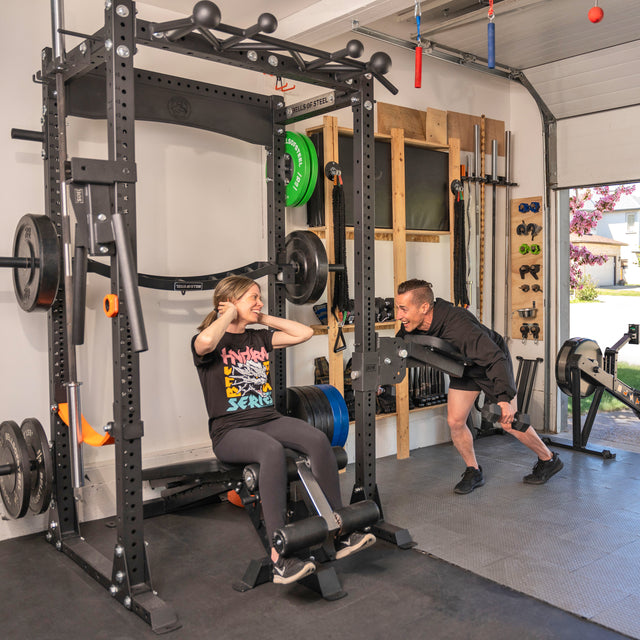 A woman uses the Bells of Steel Home Gym Builder, doing sit-ups on a weight bench while smiling at an encouraging man in a sunlit home gym with weights and power racks.