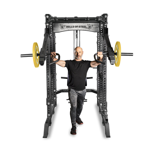 A person exercises on a Bells of Steel Lever Arms Rack Attachment. They grip handles attached to cables, with yellow weight plates on the sides, while wearing a black t-shirt and gray pants.