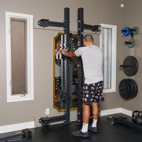 In a well-lit home gym, a person in a gray shirt and patterned shorts adjusts the Bells of Steel Manticore Folding Half Rack - Prebuilt (3" x 3", 1" Holes), with barbells and weight plates stored nearby.