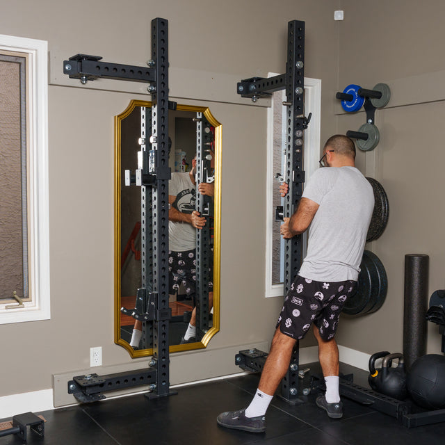 A man in a gray shirt and patterned shorts adjusts the Bells of Steel Manticore Folding Half Rack (Prebuilt, 3"x3", 1" Holes) in a home gym with weights, kettlebells, and a gold-framed mirror on the wall.