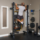 A bearded man in a gray shirt and black shorts does a pull-up on the Bells of Steel Manticore Folding Half Rack (3" x 3", 1" holes) in a home gym surrounded by weights, a mirror, and other gym equipment.