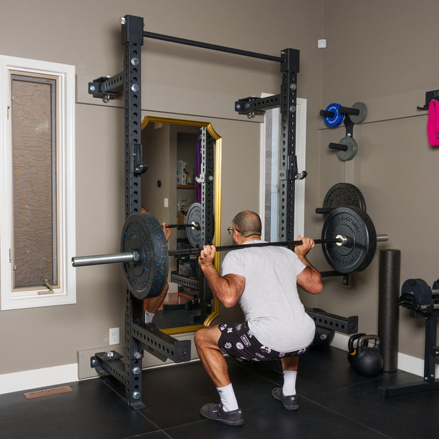 A person in a gray shirt and shorts performs a barbell squat in a home gym, facing a mirror. A Bells of Steel Manticore Folding Half Rack (3"x3", 1" holes) and various weights and gym equipment are visible around the room.