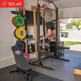 A man uses the Bells of Steel Home Gym Builder power rack in his garage gym, with barbell, bench, and weight plates nearby; a "10% off" sale banner is shown in the upper corner.