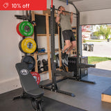 A man uses the Bells of Steel Home Gym Builder for pull-ups in his garage gym, with a weight plate set and bench close by. A red "10% off" banner appears in the corner as sunlight streams through the open door.