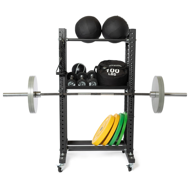 A weightlifting rack featuring a barbell loaded with weights functions as a comprehensive training setup. Above it are medicine balls and kettlebells, while a 100 lbs sandbag rests nearby. Colored weight plates are neatly arranged on the bottom shelf, with Bells of Steel Canada Utility Hooks providing additional gym storage. The rack is equipped with wheels for easy mobility.