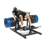 A person in a black t-shirt and shorts uses a leg press machine with large blue and black plates next to a Bells of Steel Hip Thrust Machine, lying back and pushing against the platform with their feet.