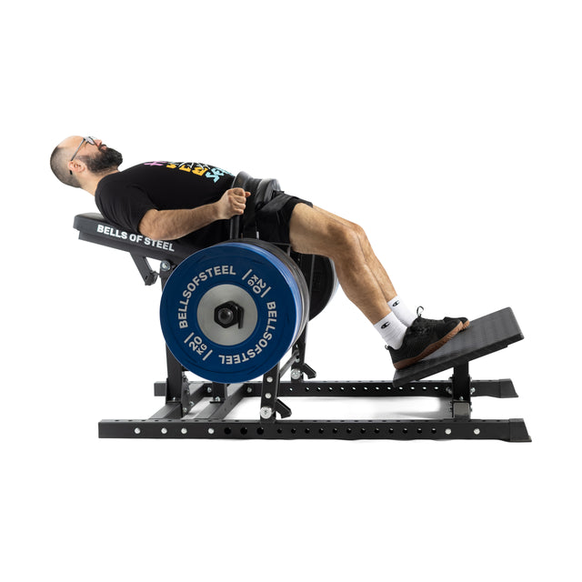 A bearded individual uses a Bells of Steel Hip Thrust Machine with an adjustable belt, leaning back on a padded bench, feet secured on a platform to exercise their lower body. Blue weights labeled "Bells of Steel" add to the workout intensity.