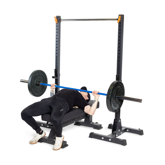 Dressed in gym attire, a person grips the Bells of Steel Multi-Purpose Olympic Barbell – The Utility Bar with determination, lying on a bench press. Loaded with weights, it forms part of a weightlifting station as they prepare for the lift.