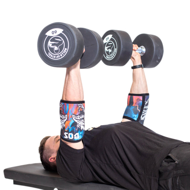 A person is lying on a gym bench performing a chest press with large, black dumbbells. They are wearing colorful compression elbow sleeves from Bells of Steel and a black shirt, with the number 505 visible on one sleeve. The plain white background emphasizes their focus and support in every rep.