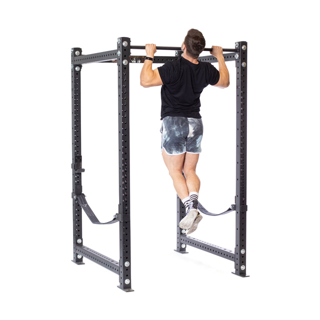 A person demonstrates a pull-up on the Bells of Steel Hydra Four Post Power Rack Builder, which features a freestanding bar. They're wearing a black shirt, tie-dye shorts, and sports shoes. The sturdy metal station is designed with visible bolts and adjustable supports for enhanced stability.