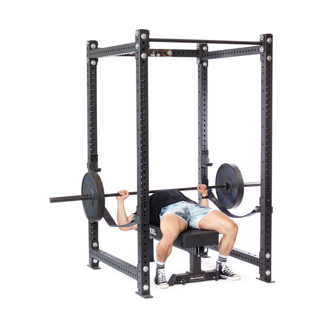 Inside a Bells of Steel Hydra Four Post Power Rack Builder, a person is performing a bench press, lifting a barbell with weights. Clad in shorts and sneakers, they lie confidently on the bench beneath the barbell, fully utilizing the secure setup of the modular components.