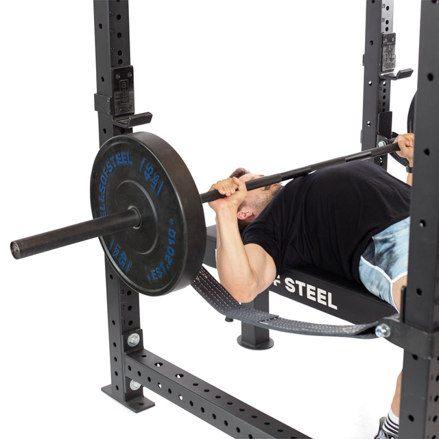 A person wearing a black shirt and blue shorts is lying on a bench, performing a bench press in a Bells of Steel Hydra Four Post Power Rack - Prebuilt (3" x 3", ⅝" Holes). They are lifting a barbell loaded with weights. The black rack, marked with white text, features modular components for compatible attachments.