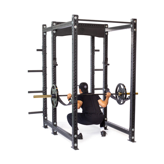 Dressed in black workout clothes, a person performs a squat inside the Hydra Six Post Power Rack Builder by Bells of Steel, capable of holding 1,000 pounds. The sturdy gym equipment features modular components like multiple support beams and storage pegs for additional weights.