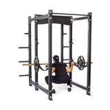 Dressed in black workout clothes, a person performs a squat inside the Hydra Six Post Power Rack - Prebuilt by Bells of Steel, lifting a barbell with weight plates. The robust power rack, featuring 3" x 3" modular components and ⅝" adjustment holes, frames the lifter's back as they face away from the camera.