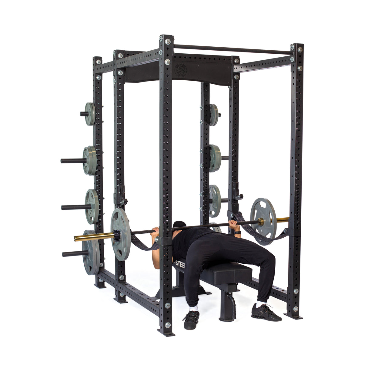 Dressed in black workout gear, an individual bench presses inside a Bells of Steel Hydra Six Post Power Rack Builder (3" x 3", ⅝" Holes) with a 1,000-pound capacity. The modular rack includes multiple weight holders and stores extra plates on the sides.