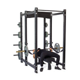 Dressed in black workout gear, an individual bench presses inside a Bells of Steel Hydra Six Post Power Rack Builder (3" x 3", ⅝" Holes) with a 1,000-pound capacity. The modular rack includes multiple weight holders and stores extra plates on the sides.