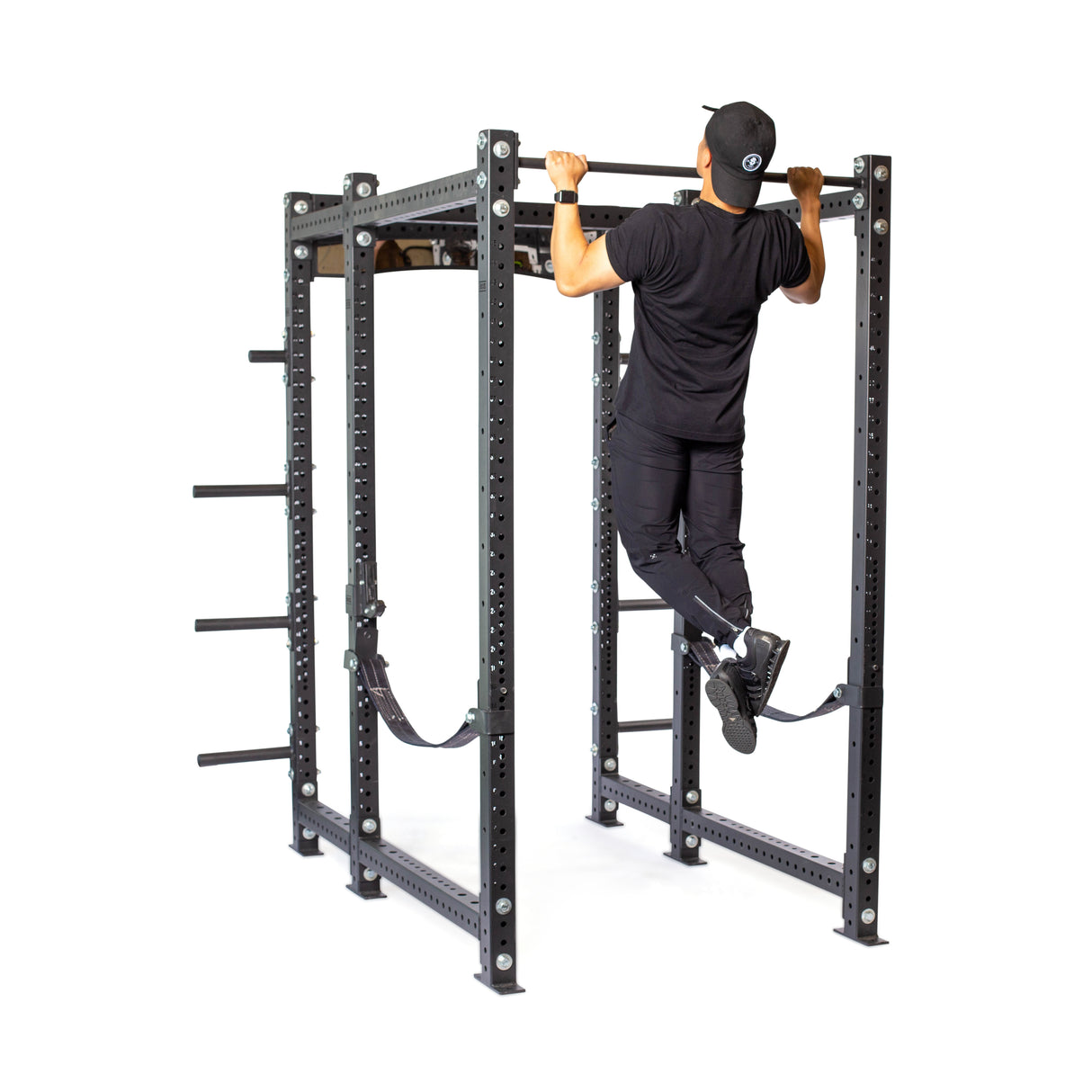 A person in a black shirt, pants, and cap performs a pull-up on the Hydra Six Post Power Rack Builder by Bells of Steel. This robust rack features multiple bars and straps, with a 1,000-pound capacity against the white background.