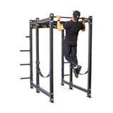 A person in a black shirt, pants, and cap performs a pull-up on the Hydra Six Post Power Rack Builder by Bells of Steel. This robust rack features multiple bars and straps, with a 1,000-pound capacity against the white background.