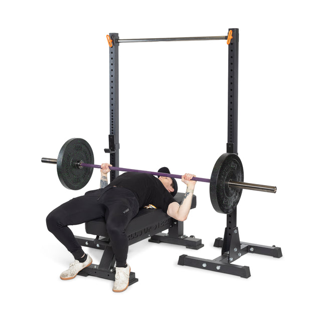 Dressed in black workout attire and white sneakers, a person performs a bench press with the Bells of Steel Multi-Purpose Olympic Barbell – The Utility Bar, which features medium-light knurling. Positioned under a sturdy rack, the setup includes a weight bench with plates.