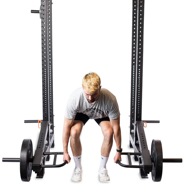 A person with short blonde hair, dressed in a gray shirt, black shorts, and white shoes, gears up to lift a barbell complete with plates. They stand in a squat position between two squat racks equipped with Bells of Steel Lever Arms Rack Attachments and an Adjustable Pull-Up Bar.