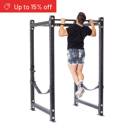A man performs a pull-up on the Bells of Steel Hydra Four Post Power Rack Builder (3" x 3", ⅝" Holes), ideal for home gyms. He wears a black shirt, tie-dye shorts, and striped socks. A red "Up to 15% off" banner appears on a plain white background.