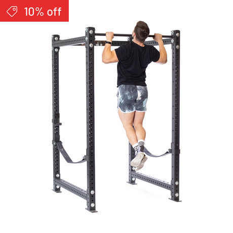 A man in a black shirt and tie-dye shorts does a pull-up on the Bells of Steel Hydra Four Post Power Rack Builder (3" x 3", ⅝" Holes). A red banner in the top left corner shows “10% off.” Perfect for any home gym.