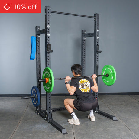 A person squats with a loaded barbell on their shoulders inside the Bells of Steel Hydra Half Rack Builder (3" x 3", ⅝" Holes), ideal for home gyms. Green and blue plates are on the bar, and a “10% off” badge is visible in a gym setting.