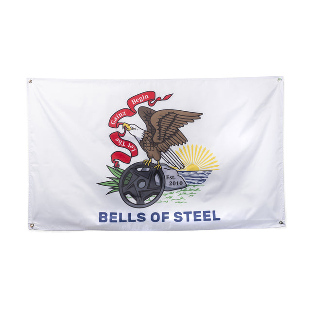 Illustration of an eagle perched on a weight plate with red ribbons featuring the text "Begin the Gainz." A sun rises behind, and the phrase "BELLS OF STEEL" enhances this striking flag from the product line by Bells of Steel. The words "Est. 2010" are displayed near the sunlit horizon, making it perfect for gym decor.