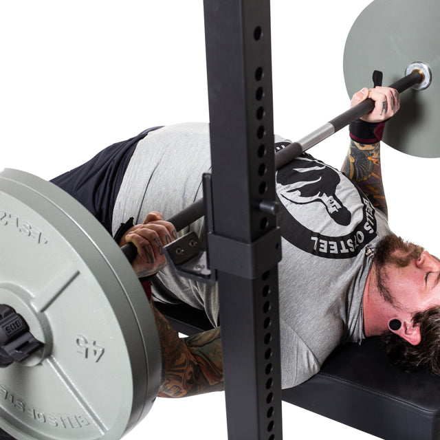 A tattooed individual, wearing a light gray T-shirt, is demonstrating strength and focus at the gym while doing a bench press using the Bells of Steel Powerlifting Bar loaded with large weights.