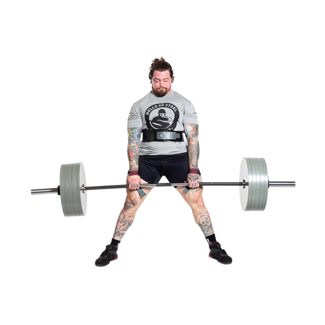 A person with tattoos is deadlifting a Bells of Steel Powerlifting Bar loaded with large weights. Wearing a gray t-shirt and black shorts, they focus intently on the lift, secured by a weightlifting belt around their waist.