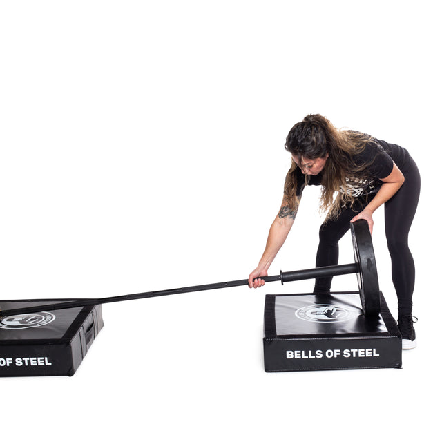 A person adjusts a weightlifting bar on a floor-mounted landmine attachment, surrounded by two Bells of Steel Deadlift Pads, while wearing athletic clothing and looking focused.