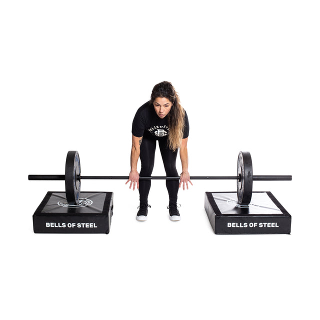 A person in workout attire lifts a barbell with weights on two square platforms labeled "Bells of Steel," featuring Deadlift Pads (Pair) that absorb the impact, set against a plain white background.