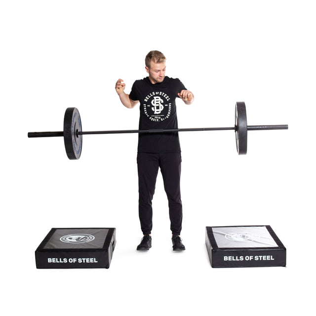 A person in a "Bells of Steel" t-shirt balances a barbell on their index fingers between Bells of Steel Deadlift Pads. The pads are designed for deadlifting and branded with the same logo. The scene is set against a plain white background.