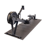 A black indoor rowing machine is placed on a Bells of Steel Cardio Machine Floor Mat, with a flywheel, sliding seat, footrests, and a monitor for workout tracking. The setup is pictured against a white background.