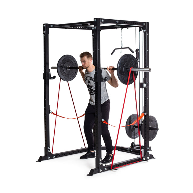 A person is performing a squat with a barbell inside a black power rack, capturing the essence of powerlifting. The rack is equipped with weight plates and 41" Fabric Non-Slip Resistance Bands from Bells of Steel. They are dressed in a gray T-shirt and black pants, fully concentrated on their exercise.