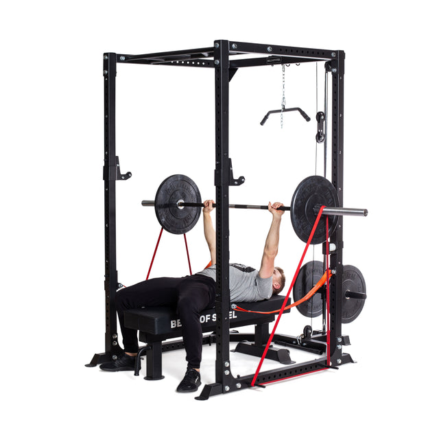 Lying on a bench within the power rack, they lift a barbell loaded with weights, while Bells of Steel's Fabric Non-Slip Resistance Bands (41") increase the intensity. Surrounded by other gym attachments, this setup evokes powerlifting training and stands out against the plain white background.