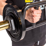 A person wearing a black shirt and shorts grips a black trap bar loaded with a "Bells of Steel" weight plate, secured by Bells of Steel Magnetic Clamp Collars, ready for lifting.