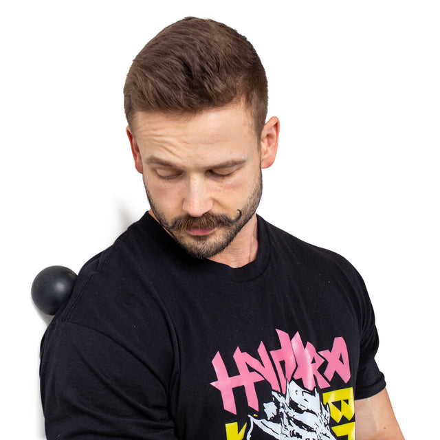 A mustached man in a black t-shirt with colorful graphics uses the Bells of Steel Massage Balls against a wall for upper back muscle relief, making it his go-to tool for easing tension.