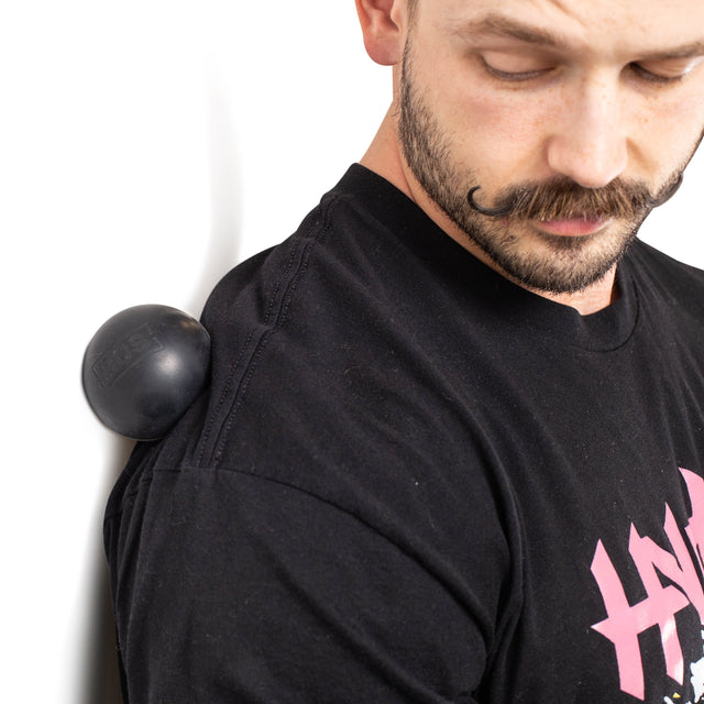 A person in a black T-shirt with a pink design and a distinctive curled mustache uses Bells of Steel Massage Balls for muscle relief between their shoulder and the wall, focusing intently while looking down.