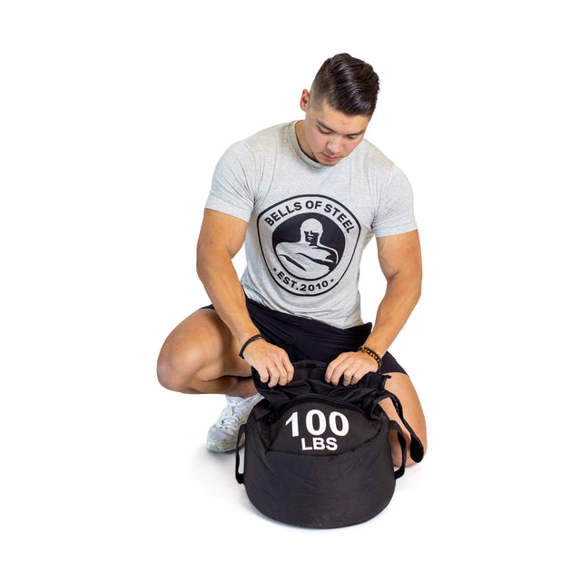 A man in a light gray T-shirt, featuring a circular logo, crouches to open a black Bells of Steel Fitness Sandbag marked "100 LBS," highlighting its durable Condura construction. He pairs this look with black shorts and white sneakers against a plain white background, ready for sandbag workouts.