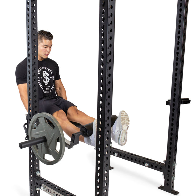 In his home gym, a man wearing a black t-shirt and shorts effectively performs leg exercises using the Bells of Steel Leg Curl/Leg Extension Rack Attachment with weight plates.